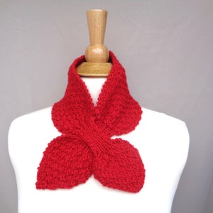 Könnte beinhalten: Ein leuchtend roter Strickschal, präsentiert auf einer weißen Schaufensterpuppe. Der Schal hat eine einzigartige Form mit einem zentralen Knoten und ausgestellten Enden. Die Textur des Stricks ist deutlich sichtbar. Der Hintergrund ist grau.