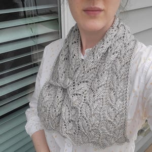 May include: A light grey knitted cowl scarf featuring a leaf pattern. The scarf is made from a soft, textured yarn and has a scalloped edge. The cowl is worn around the neck and is a stylish accessory.