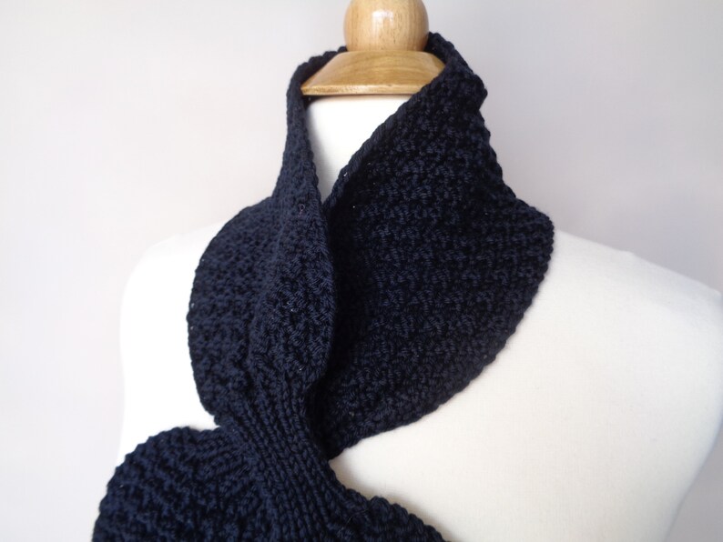 May include: A black knitted infinity scarf with a textured pattern. The scarf is draped over a white mannequin.