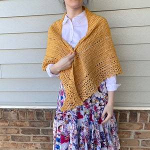 May include: A yellow crochet shawl with a scalloped edge. The shawl is draped over a person's shoulders and is being held in place by one hand.