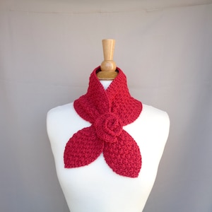 May include: A red crocheted scarf with a floral design. The scarf is displayed on a white mannequin. The scarf has a textured appearance and features a large, three-petal flower in the center.