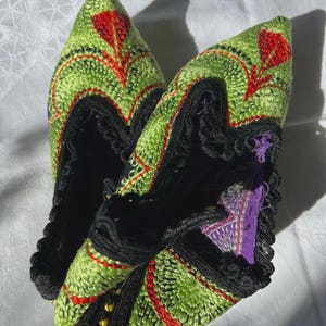 Op de afbeelding: Geborduurde pantoffels met een puntige neus. De pantoffels hebben een groene achtergrond met rode en zwarte borduursels. De binnenkant is zwart met een paarse accent.