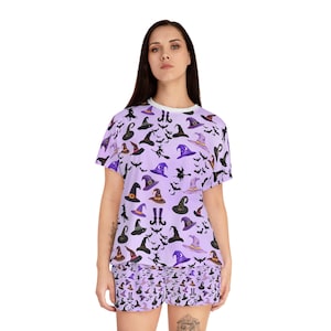 May include: A lilac-colored pajama set featuring a repeating pattern of witch hats, bats, and witch silhouettes. The short-sleeved top and matching shorts have a white trim. The witch hats are in various colors, including black, purple, and brown.