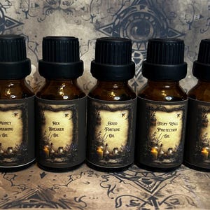 Może przedstawiać: Pięć butelek z brązowego szkła z czarnymi zakrętkami, każda oznaczona inną nazwą: Money Drawing Oil, Hex Breaker Oil, Good Fortune Oil, Fiery Wall Protection Oil i Pathway Opening Oil. Etykiety mają ciemną obwódkę z motywem świec i kwiatów.