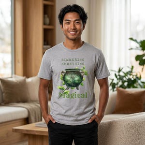 May include: A gray t-shirt featuring a green cauldron design with the words "Simmering Something Magical." The cauldron is detailed with green and gold accents, surrounded by green leaves. The shirt is worn by a person.