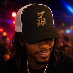 May include: A black and white trucker cap featuring the number 718 embroidered in gold on the front panel. The cap has a solid black front, a curved brim, and a white mesh back panel, providing a structured look with a classic adjustable design.