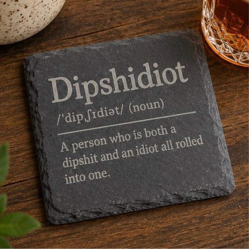 May include: A square, dark gray slate coaster with the word "Dipshidiot" engraved in a light gray font. The definition of the word is also engraved on the coaster. A decorative item for a home bar or kitchen.