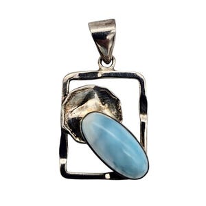 May include: A silver pendant with a rectangular frame, featuring a light blue oval gemstone and a textured silver accent. The pendant has a bail for a chain and is set against a white background.