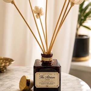 Puede incluir: Un frasco difusor de varillas de vidrio ámbar cuadrado con una etiqueta que dice The Atrium 1935 Art Deco Collection Olive Leaf & Citron White Tea Black Fig Sandalwood. Incluye varillas de madera con flores blancas y un tapón de corcho dorado.