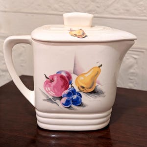 May include: Cream-colored ceramic pitcher with a lid and handle. The pitcher is decorated with a colorful illustration of fruit, including a red apple, purple plums, blue grapes, and a yellow pear. A small fruit image is on the lid.