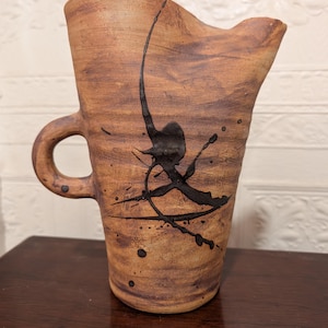 May include: Handmade ceramic pitcher with a brown and tan mottled finish. The pitcher features a handle and a spout. Black abstract paint splatters decorate the front of the pitcher. The pitcher is sitting on a dark brown surface.