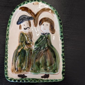 May include: A ceramic wall plaque with a glossy off-white finish featuring a hand-painted illustration of two figures in green clothing. The plaque has a scalloped green border, a brown arch shape above the figures, and a small twine loop for hanging.