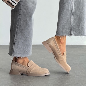 May include: Beige suede loafers with a square toe and a low, stacked heel. The shoes have a slip-on design with a strap detail across the top. The person is wearing grey jeans and holding a magazine.