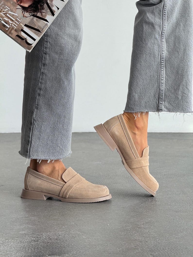 May include: Beige suede loafers with a square toe and a low, stacked heel. The shoes have a slip-on design with a strap detail across the top. The person is wearing grey jeans and holding a magazine.