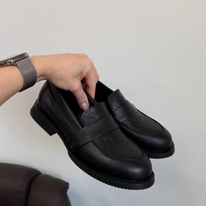 May include: A pair of black leather loafers with a textured vamp and a smooth, rounded toe. The shoes have a low, thick sole and a strap across the top. The shoes are held by a person's hand against a white background.