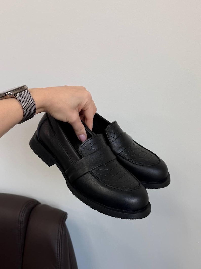 May include: A pair of black leather loafers with a textured vamp and a smooth, rounded toe. The shoes have a low, thick sole and a strap across the top. The shoes are held by a person's hand against a white background.
