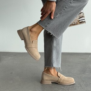 May include: Beige suede loafers with a rounded toe and a strap detail. The shoes have a low, block heel and a matching sole. The person is wearing grey jeans and a grey blazer.
