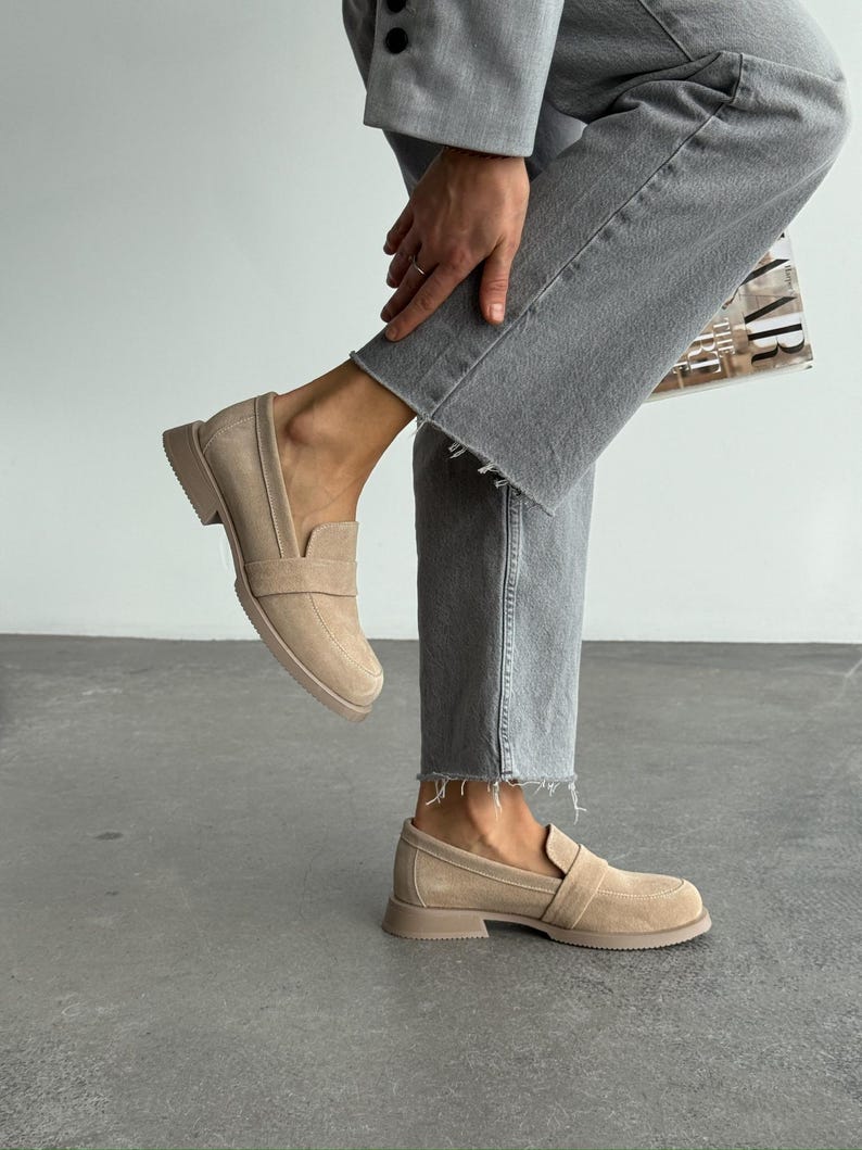 May include: Beige suede loafers with a rounded toe and a strap detail. The shoes have a low, block heel and a matching sole. The person is wearing grey jeans and a grey blazer.