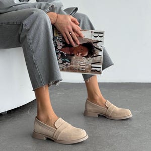 May include: Beige suede loafers with a rounded toe and a low heel. The slip-on shoes feature a strap detail. The image shows the shoes on a person wearing grey jeans. The shoes are suitable for everyday wear.
