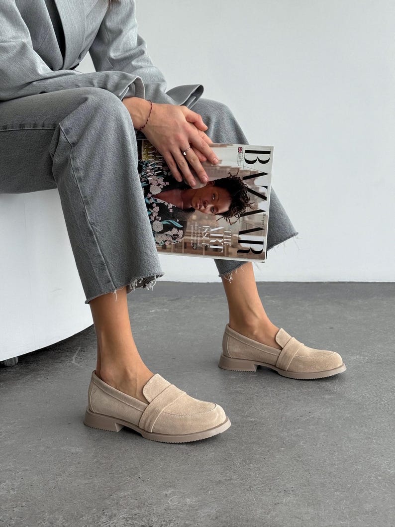 May include: Beige suede loafers with a rounded toe and a low heel. The slip-on shoes feature a strap detail. The image shows the shoes on a person wearing grey jeans. The shoes are suitable for everyday wear.