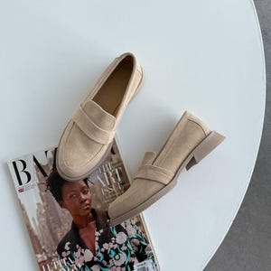 May include: Beige suede loafers with a rounded toe and a strap detail. The shoes are placed on a white surface, alongside a magazine featuring the text "BAZAAR" and "THE ART ISSUE". The shoes are approximately 25 cm long.