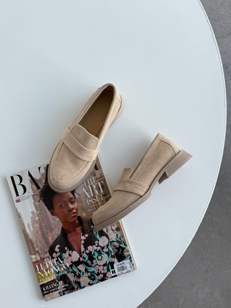 May include: Beige suede loafers with a rounded toe and a strap detail. The shoes are placed on a white surface, alongside a magazine featuring the text "BAZAAR" and "THE ART ISSUE". The shoes are approximately 25 cm long.