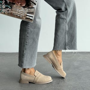 May include: Beige suede loafers with a rounded toe and a low, flat sole. The shoes have a slip-on design with a strap detail across the top. The image also shows a person wearing grey jeans and holding a magazine.