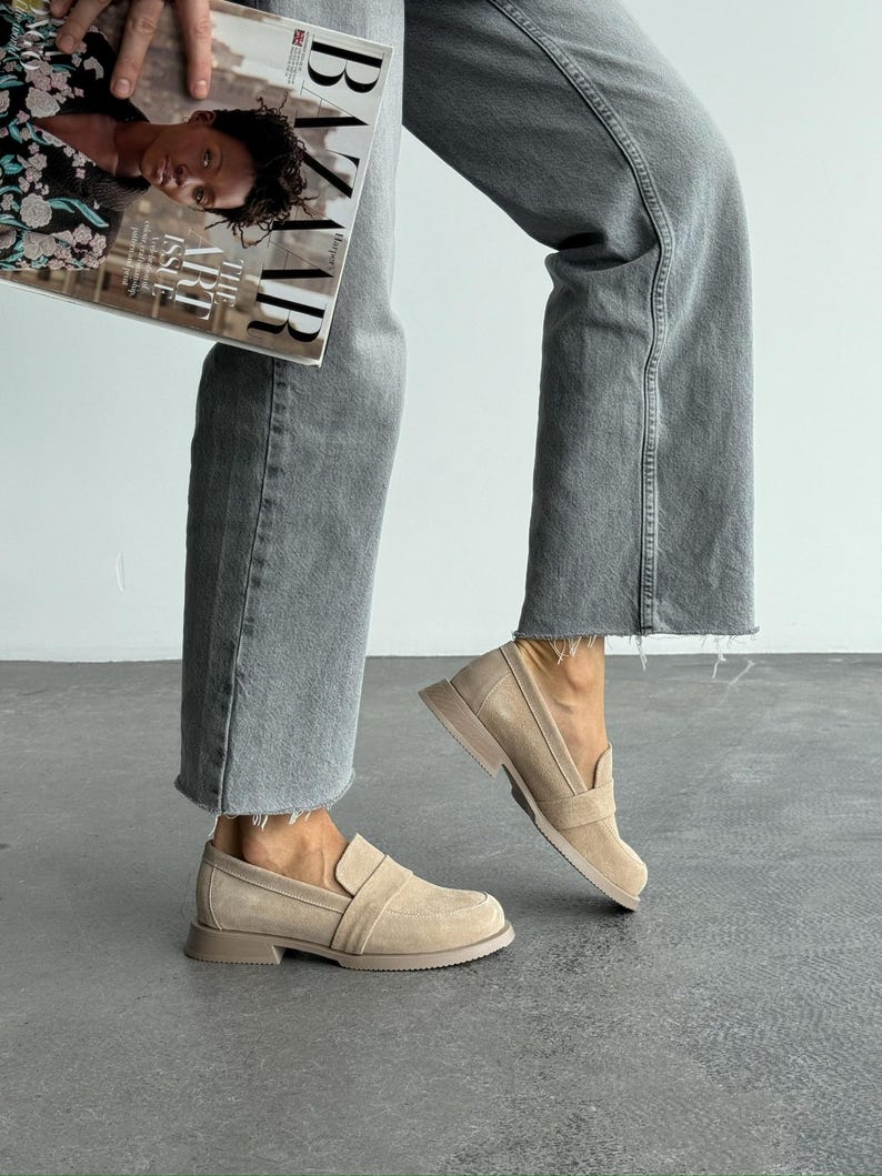 May include: Beige suede loafers with a rounded toe and a low, flat sole. The shoes have a slip-on design with a strap detail across the top. The image also shows a person wearing grey jeans and holding a magazine.