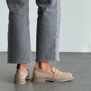 May include: Beige suede loafers with a square toe and a low, block heel. The shoes have a strap detail across the top and are worn with grey denim jeans with a raw hem. The shoes are a neutral colour.