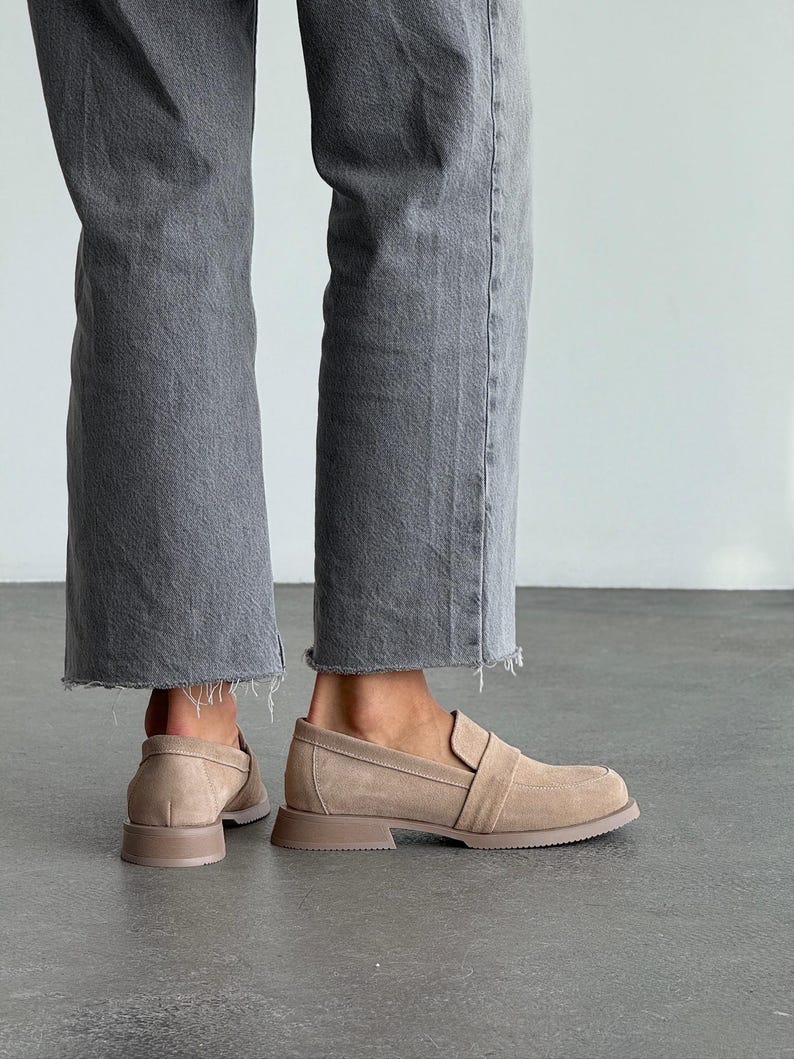 May include: Beige suede loafers with a square toe and a low, block heel. The shoes have a strap detail across the top and are worn with grey denim jeans with a raw hem. The shoes are a neutral colour.