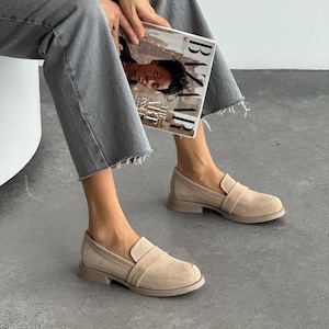 May include: Beige suede loafers with a low heel. The shoes have a rounded toe and a strap detail across the top. The soles are a matching beige colour. The person is holding a magazine.