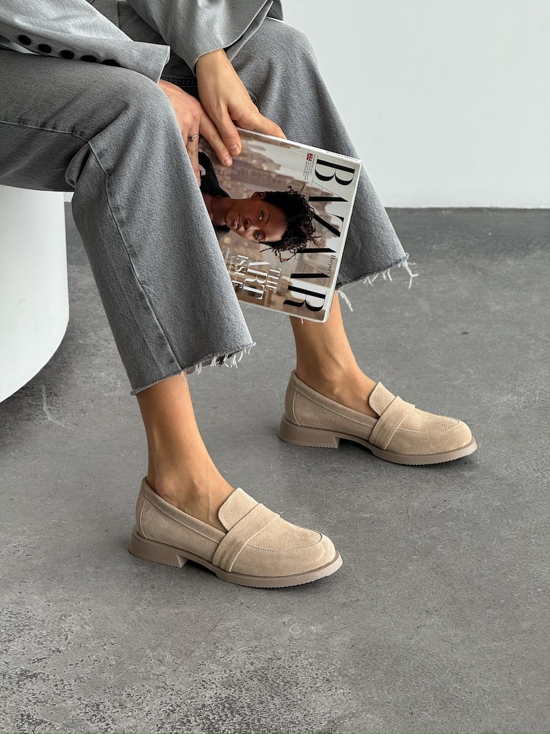 May include: Beige suede loafers with a low heel. The shoes have a rounded toe and a strap detail across the top. The soles are a matching beige colour. The person is holding a magazine.
