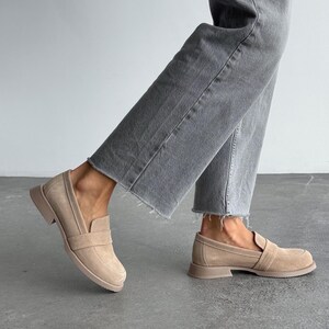 May include: Beige suede loafers with a slip-on design. The shoes feature a rounded toe and a low, stacked heel. The soles and heels are a matching beige colour. The shoes are paired with grey denim jeans.