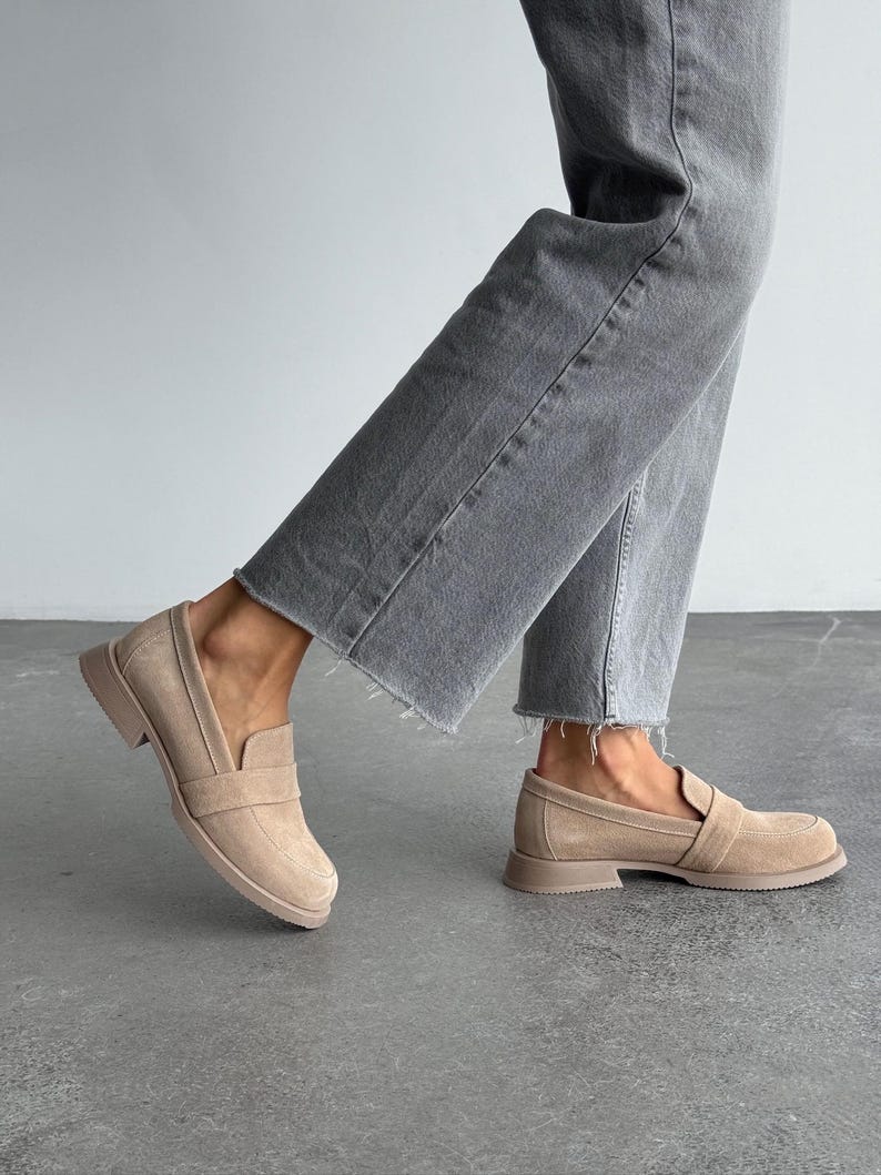 May include: Beige suede loafers with a slip-on design. The shoes feature a rounded toe and a low, stacked heel. The soles and heels are a matching beige colour. The shoes are paired with grey denim jeans.