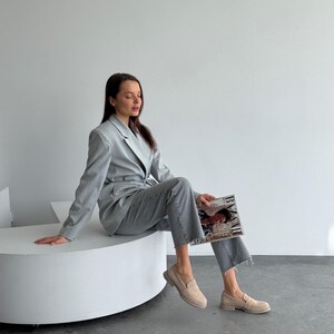 May include: A light grey suit, comprising a blazer and matching trousers, worn with beige loafers. The outfit is accessorized with a magazine. The blazer has a classic design, and the trousers have a straight leg cut. The loafers are a neutral colour.