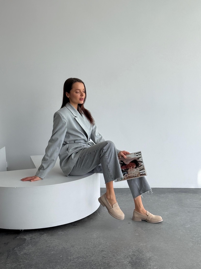 May include: A light grey suit, comprising a blazer and matching trousers, worn with beige loafers. The outfit is accessorized with a magazine. The blazer has a classic design, and the trousers have a straight leg cut. The loafers are a neutral colour.
