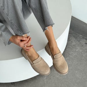 May include: Beige suede loafers with a rounded toe and a strap detail. The shoes are worn with grey denim jeans. The loafers have a low heel and a neutral colour palette. The shoes are approximately 25 cm long.