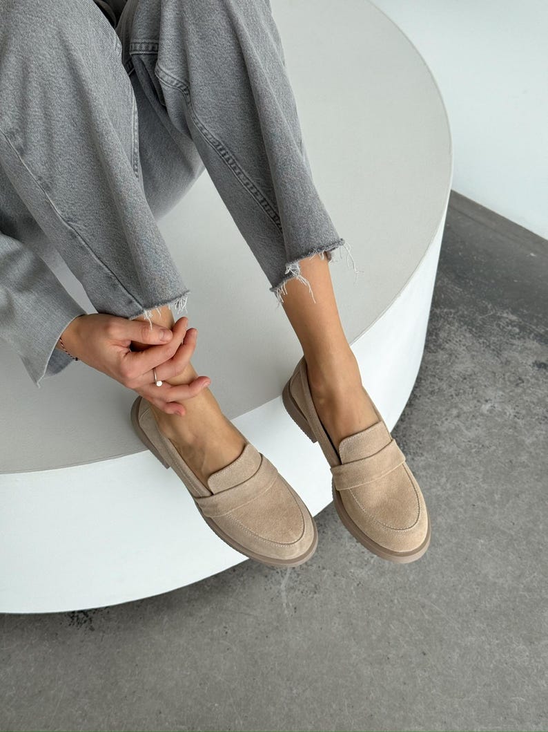 May include: Beige suede loafers with a rounded toe and a strap detail. The shoes are worn with grey denim jeans. The loafers have a low heel and a neutral colour palette. The shoes are approximately 25 cm long.