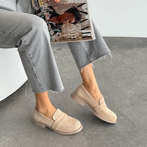 May include: Beige suede loafers with a rounded toe and a low, stacked heel. The shoes have a slip-on design with a strap detail across the top. The soles are a matching beige colour. The image also shows a person wearing grey jeans and holding a magazine.