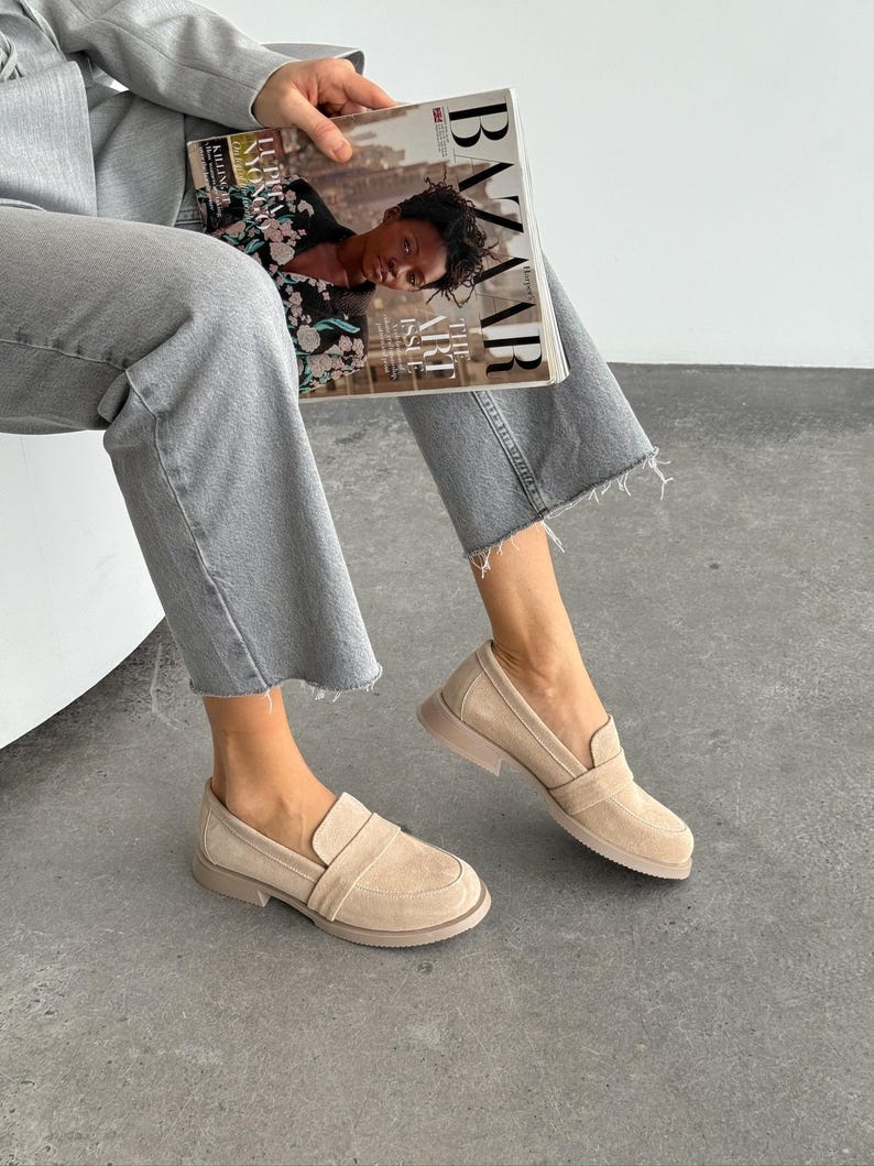 May include: Beige suede loafers with a rounded toe and a low, stacked heel. The shoes have a slip-on design with a strap detail across the top. The soles are a matching beige colour. The image also shows a person wearing grey jeans and holding a magazine.