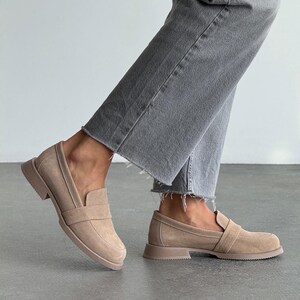 May include: Tan suede loafers with a slip-on design. The shoes feature a low, block heel and a strap detail. The shoes are worn with grey denim jeans. The shoes are approximately 2 cm high.