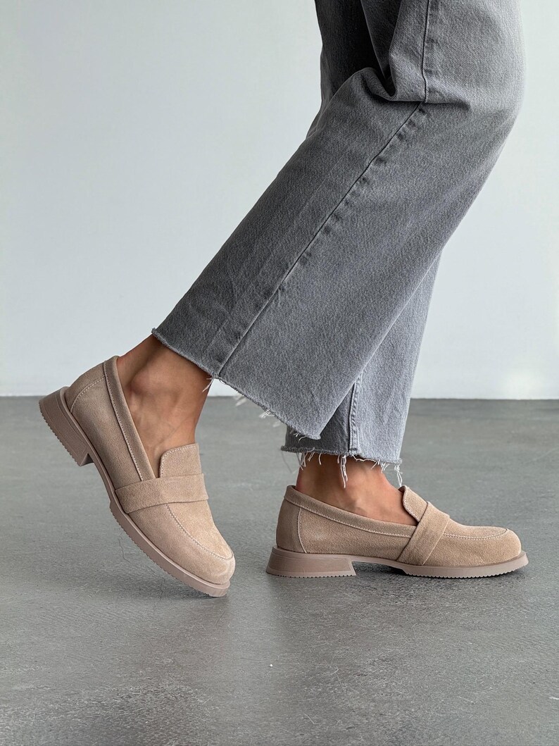 May include: Tan suede loafers with a slip-on design. The shoes feature a low, block heel and a strap detail. The shoes are worn with grey denim jeans. The shoes are approximately 2 cm high.