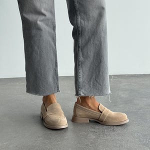 May include: Beige suede loafers with a rounded toe and a low, block heel. The slip-on shoes have a strap across the top. The image shows the shoes from a low angle, with the wearer's legs in grey denim jeans.