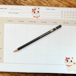 May include: A weekly planner with a cat theme, featuring a grid for each day, a habit tracker, and space for notes. A black pencil rests on the planner. The planner has a light beige background with cat illustrations.