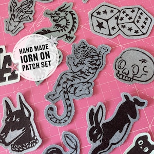 May include: A collection of iron-on patches in various shapes and designs. The patches include a tiger, skull, dice, dragon, and a rabbit. The patches are made of denim fabric with black designs. The text "HAND MADE IRON ON PATCH SET" is in a circle.