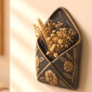Puede incluir: Una maceta negra con forma de sobre montada en la pared con detalles florales y espigas de trigo doradas. La maceta está llena de flores amarillas secas y trigo. La maceta está montada en una pared beige.