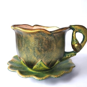May include: A decorative ceramic teacup and saucer set. The teacup is shaped like a flower with a gold and green finish. The saucer is leaf-shaped and green. The handle has a unique, curved design.