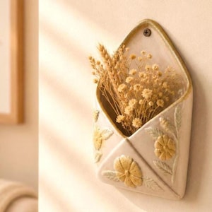 Puede incluir: Una maceta de cerámica en forma de sobre, de color crema, montada en la pared. La maceta está decorada con diseños florales y de hojas en relieve en amarillo y dorado. Está llena de flores secas y espigas de trigo. Un acento decorativo para la decoración del hogar.