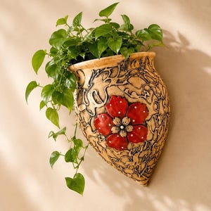 Puede incluir: Una maceta de pared con una superficie beige texturizada que presenta un patrón negro similar a una enredadera y una flor roja prominente con puntos blancos en el centro. Una planta verde de hojas crece desde la parte superior del recipiente.