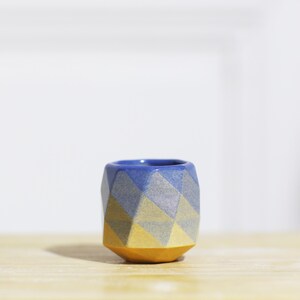 Op de afbeelding: Een kleine, gefacetteerde keramische beker met een geometrisch ontwerp. De beker heeft een blauw interieur, met een patroon van blauwe, grijze en gele facetten aan de buitenkant. De basis is effen geel. De beker staat op een houten oppervlak.